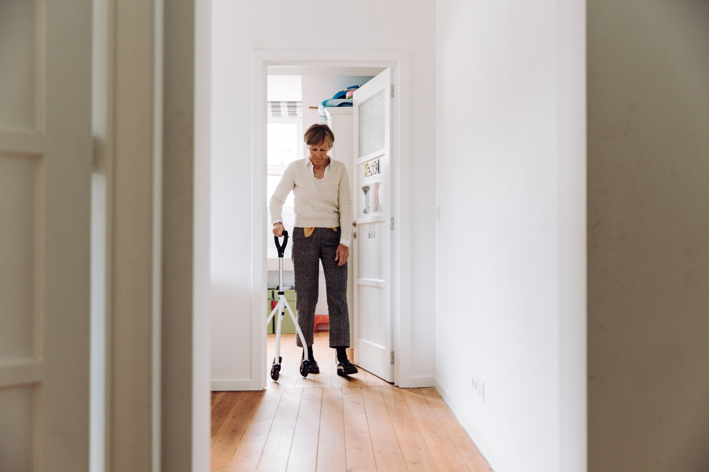 An older woman walks through a bright hallway using a wheeled walking aid.