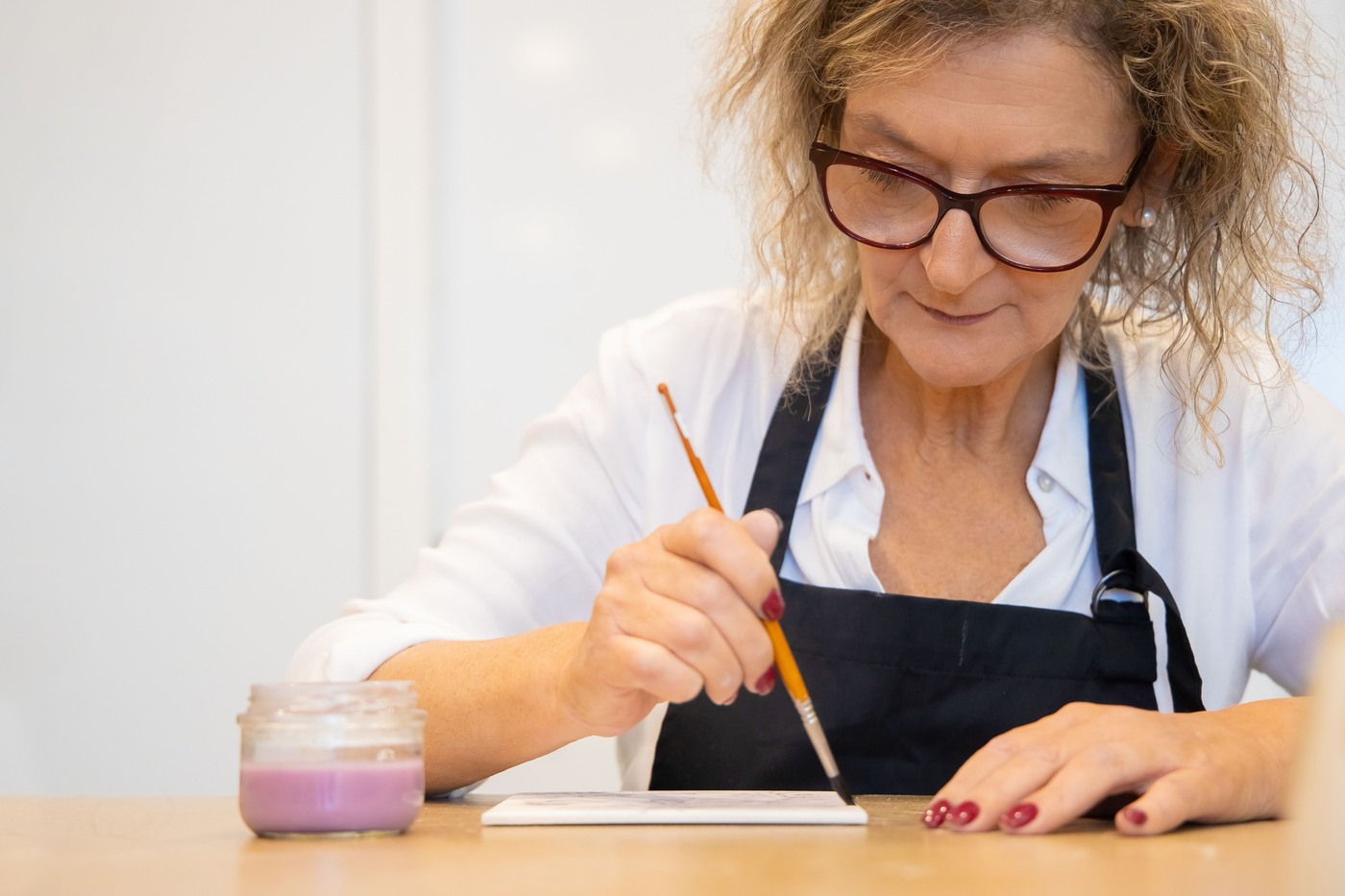 An older woman concentrates on painting a ceramic tile at a table.