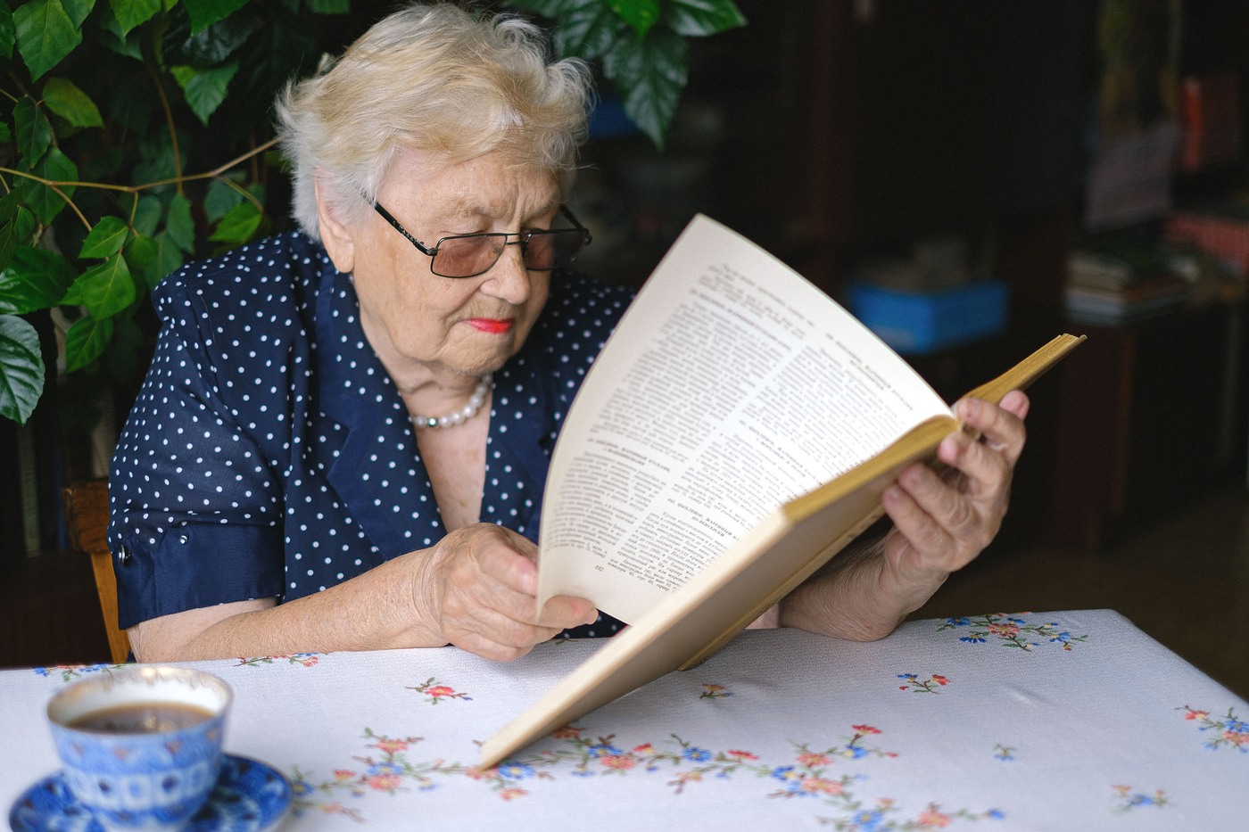 A woman in her seventies reads a paperback at her kitchen table in morning light.